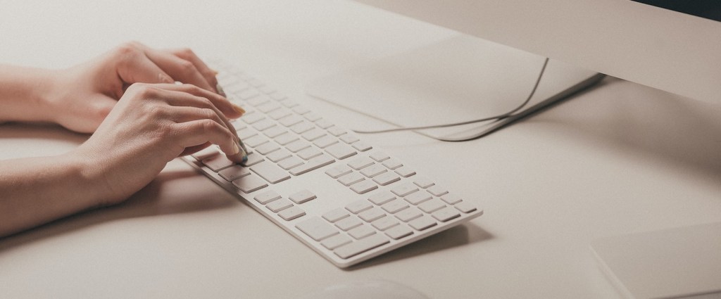 Pix Saque Pix Troco Banco Central Consulta Pública: fotografia de uma pessoa digitando com as duas mãos em um teclado branco. Ao lado, um mouse branco e, na frente, a tela de um computador (Créditos: Ilya Pavlov)