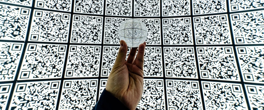 Pix cobrança para pagamentos com vencimento: fotografia de uma mão segurando um globo de vidro num fundo cheio de QR codes repetidos (Crédito Mitya Ivanov)