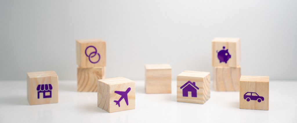 Imagem de vários cubinhos de madeira espalhados em uma superfície branca, com carro, cofre de porquinho, avião, casa, loja e dois círculos.