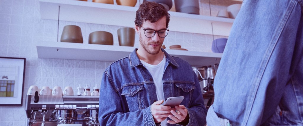 cobrança clientes pix: homem segura o celular em um café aparentemente gerando uma cobrança para uma cliente que está de costas de jaqueta jeans