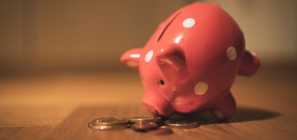 um cofrinho de porquinho rosa, com bolinhas brancas, em cima de algumas moedas. Foto: Andre Taissin