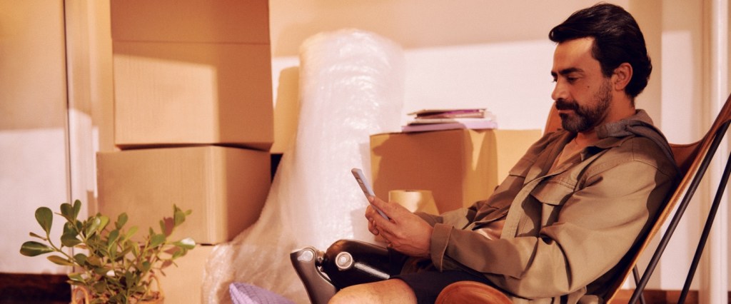 homem de meia idade com o celular na mão com caixas próximas a ele que se assemelham a uma mudança sentado numa cadeira de descanso
