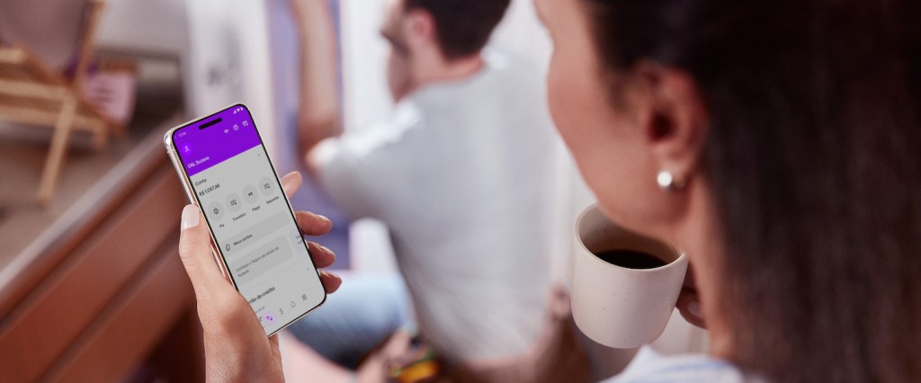 Imagem de uma mulher segurando uma xícara de café em uma mão e o celular na outra. Na tela do aparelho, o app do Nubank. No fundo na imagem tem um homem abaixado, consertando algo na parede.
