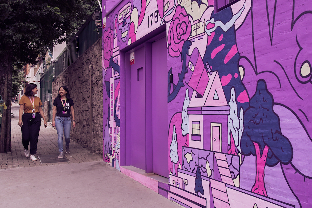 Fachada de uma casa toda grafitada com imagens roxas. Duas mulheres vêm pela calçada conversando e sorrindo.