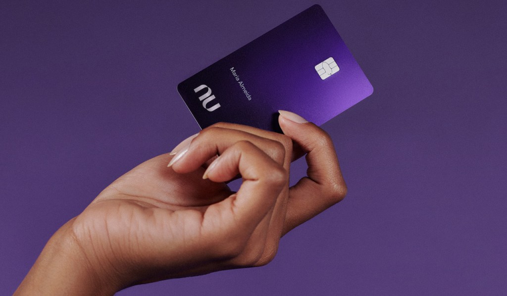 Fundos Nu Ultravioleta: fotografia de uma mão segurando o cartão Nubank Ultravioleta com um fundo roxo escuro atrás