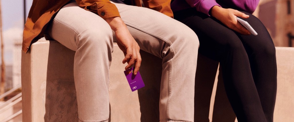 Foto mostra as pernas de duas pessoas sentadas em uma mureta, uma com calça clara, a outra com calça escura - e uma delas segura o cartão Nubank roxo