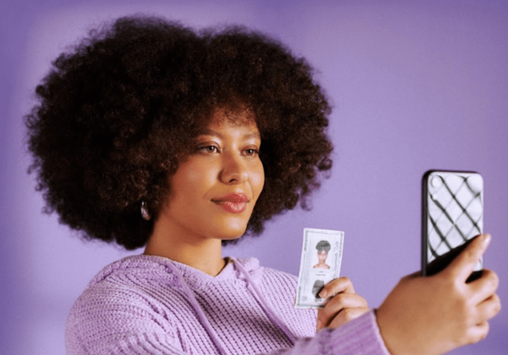 mulher segurando o documento pessoal de frente para o celular, para tirar uma selfie. O fundo é roxo