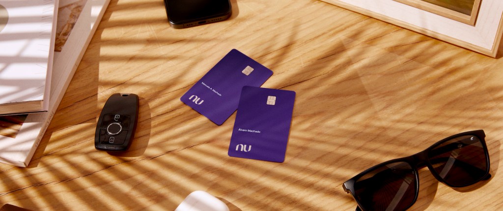 Foto mostra dois cartões Ultravioleta em cima de uma mesa de madeira clara. Ao lado deles há um óculos escuros, uma chave de carro, um celular e um fone de ouvido.