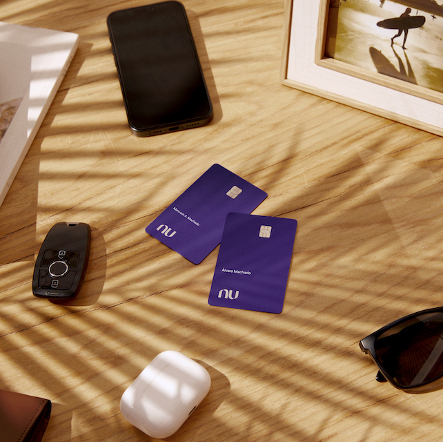 Foto mostra dois cartões Ultravioleta em cima de uma mesa de madeira clara. Ao lado deles há um óculos escuros, uma chave de carro, um celular e um fone de ouvido.