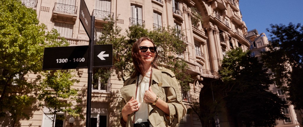 Foto mostra uma mulher branca de cabelos lisos e castanhos, com óculos escuros e uma capa bege e camisa branca. Ela está em uma viagem internacional.