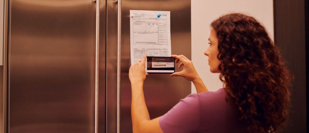 Mulher de cabelos longos e en aracolados vestindo uma camiseta roxa aponta o celular para um boleto que está fixado na porta da geladeira