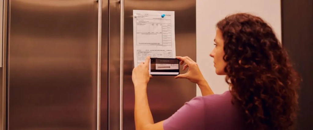 Mulher de cabelos longos e encaracolados vestindo uma camiseta roxa aponta o celular para um boleto que está fixado na porta da geladeira