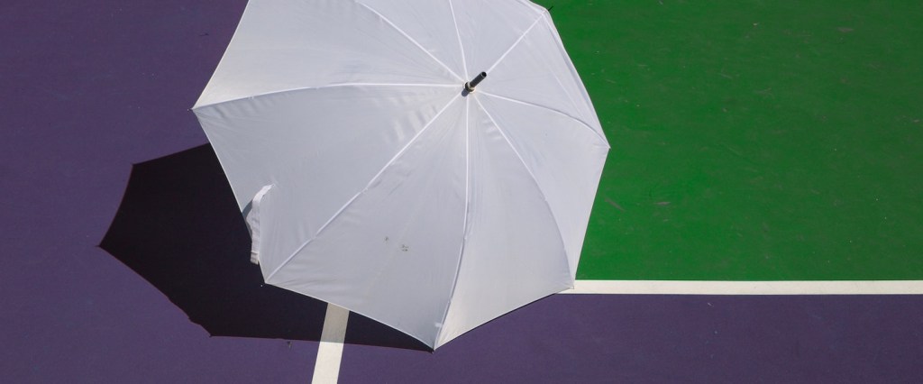 Quarta parcela auxílio emergencial 2021 nascidos outubro: fotografia de um guarda-chuva branco sobre uma quadra esportiva roxa e verde com a sombra da rede de basquete ao fundo. Créditos da imagem: Marra
