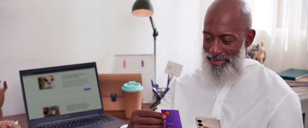 Imagem de um homem negro segurando um cartão Nubank numa mão e um celular na outra. Ele está sentado e atrás, numa mesa, tem um notebook aberto na página do blog do Nubank.