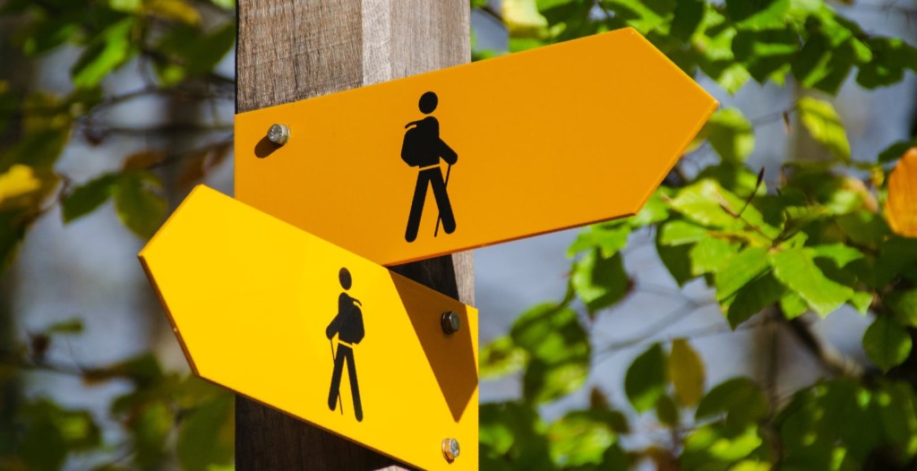 Foto de um poste com duas placas de trânsito contendo a sinalização de uma pessoa caminhando. A imagem ilustra os diversos caminhos possíveis para escolher fundo de investimento, discussão abordada neste conteúdo.