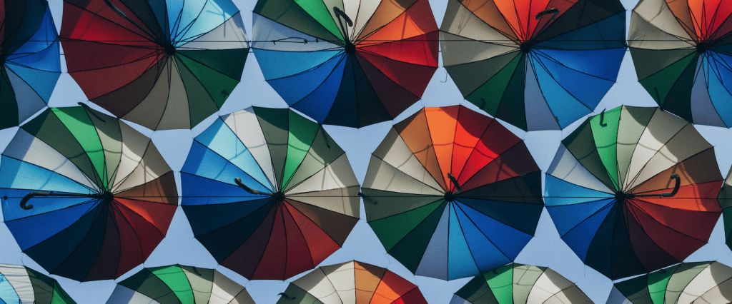 Auxílio emergencial 2021: guarda-chuvas abertos vistos de baixo. Todos com faixas coloridas com céu azul ao fundo. Créditos: Marjan Blan, Unsplash.