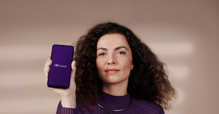 Foto de uma mulher de cabelo cacheado usando um casaco roxo. Ela está segurando um celular com a tela inicial do app Nu invest