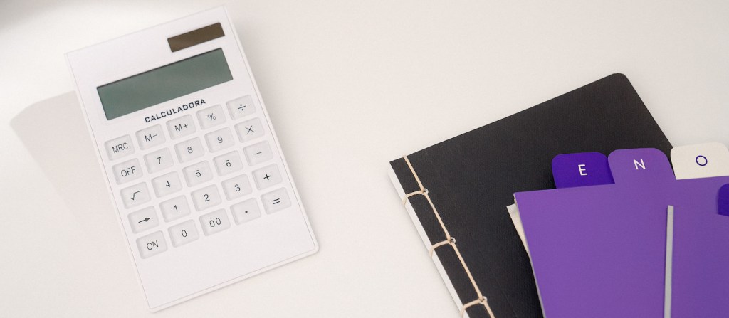 Imagem de uma calculadora branca ao lado de uma pasta preta e fichas em tons de lilás e roxo.