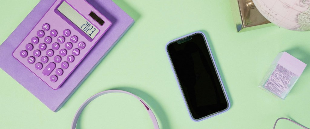 Imagem de uma calculadora lilás, com os números 2023 na tela. Um fone de ouvido grande lilás, um celular com a tela preta, uma caixinha de clip lilás. Tudo em uma superfície verde claro.