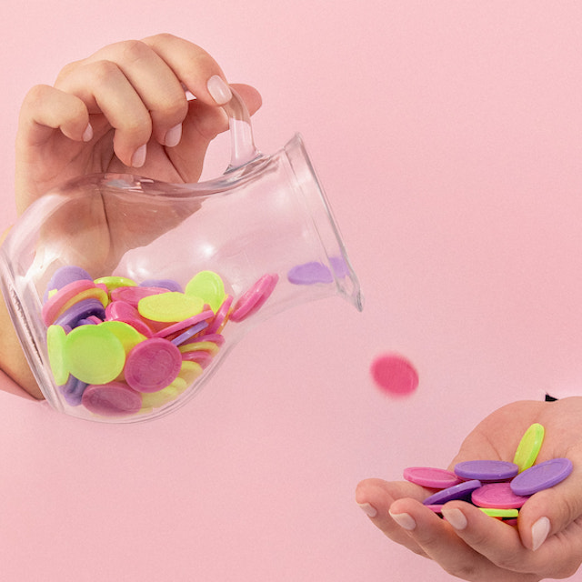 Imagem de duas mãos onde uma derruba uma jarra com moedas coloridas e a outra segura as moedas que caem da jarra. As mãos estão saindo de um fundo de papel rosa claro.