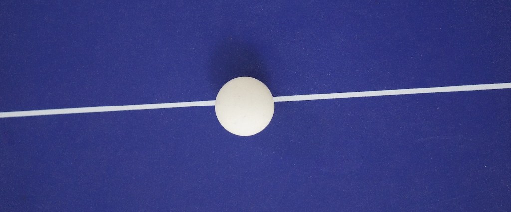 Stablecoin: fotografia de uma bolinha de ping pong branca sobre uma linha branca que cruza da direita pra esquerda no fundo azul. Créditos da imagem: Kiran Ck