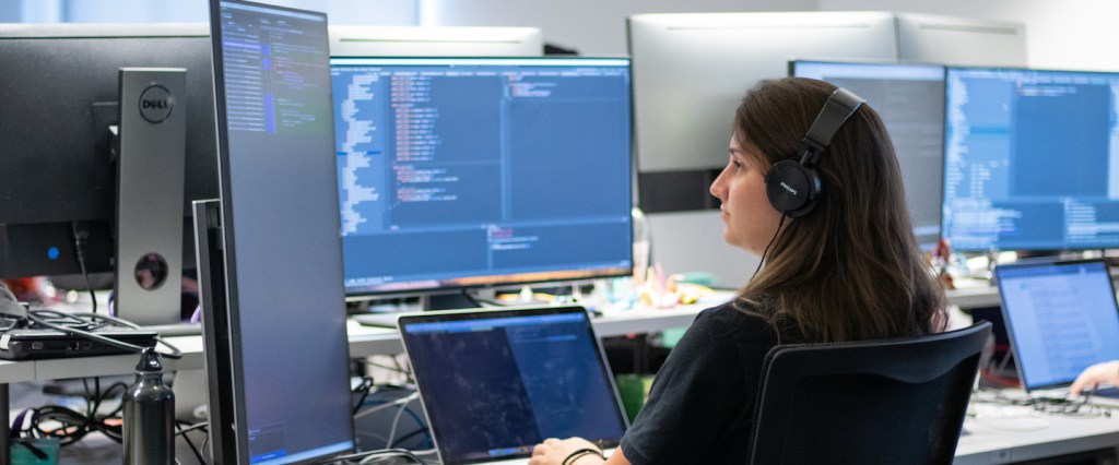 Engenharia de software Nubank: uma mulher com fones de ouvido programando. Ela está com um notebook na mesa e usa dois monitores auxiliares.