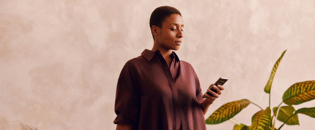 Mulher negra com cabelos curtos e vestindo uma camisa marrom observa o celular.