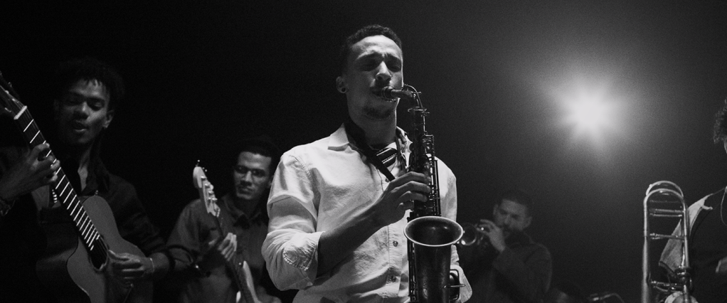 Propaganda Nubank, a história de Lucas: fotografia em preto e branco de Lucas tocando saxofone com os olhos fechados. Ao fundo, outras pessoas tocam outros instrumentos musicais, como uma guitarra e um clarinete.