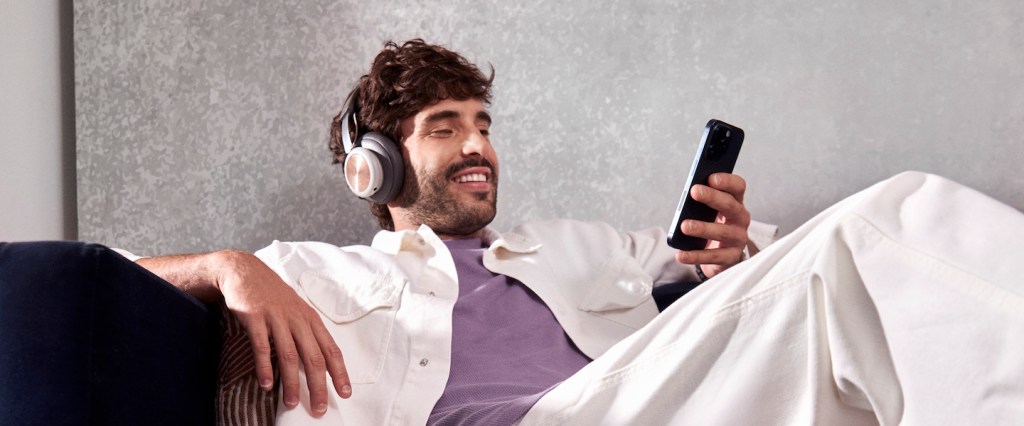 Um homem branco sentado confortavelmente em um sofá, sorrindo enquanto usa um headset e olha para o celular em uma de suas mãos.