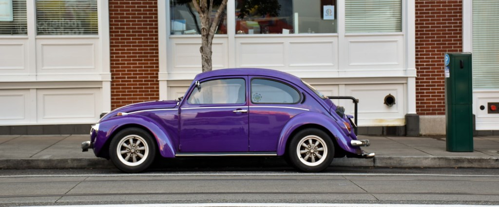 O que é DUT? Fusca roxo estacionado, ao fundo, tronco fino de uma árvore e prédio com paredes brancas e tijolos marrons. Créditos: Elena Kuchko - Unsplash