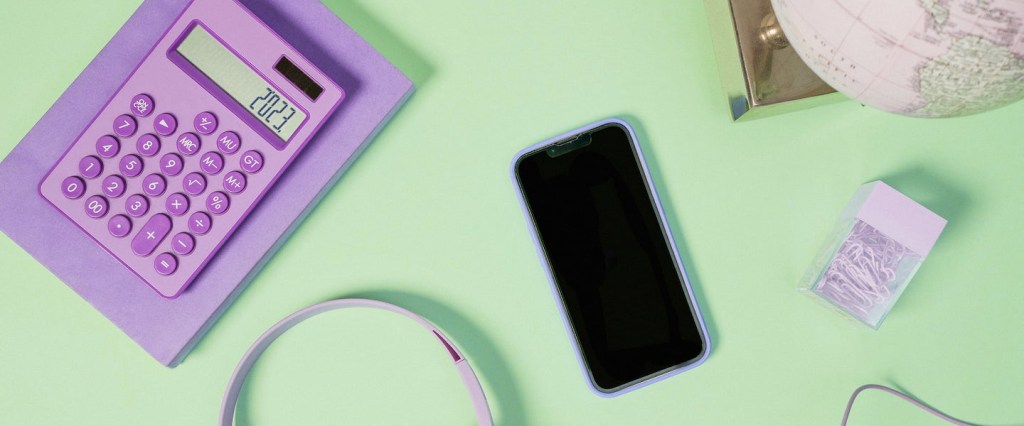 Imagem de uma calculadora lilás, com os números 2023 na tela, um celular desligado no meio, uma caixinha de clip, um globo terrestre e um headphone. Tudo numa superfície verde clara.