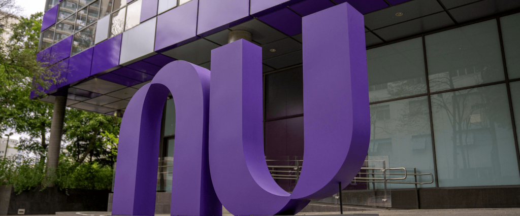 Logo roxo do Nubank na frente da sede do escritório em São Paulo. Ao fundo, portas de vidro.