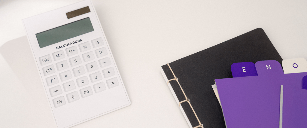 Como emitir DARF: imagem de uma calculadora branca ao lado de um caderno preto e de etiquetas roxas sobre uma mesa de escritório branca.