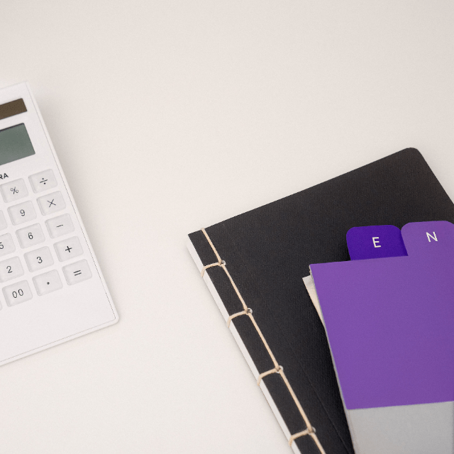 Como emitir DARF: imagem de uma calculadora branca ao lado de um caderno preto e de etiquetas roxas sobre uma mesa de escritório branca.