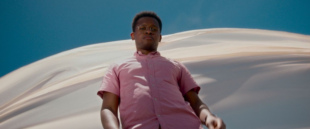 Na imagem aparece um homem negro com uma camisa em tom roxo claro olhando para baixo na direção da câmera, e atrás dele um pano esvoaçante branco com céu azulado