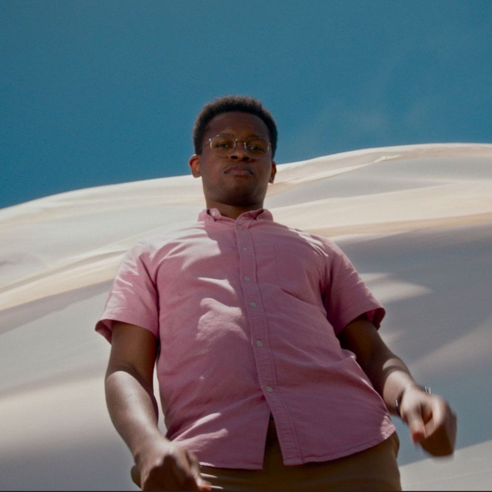 Na imagem aparece um homem negro com uma camisa em tom roxo claro olhando para baixo na direção da câmera, e atrás dele um pano esvoaçante branco com céu azulado