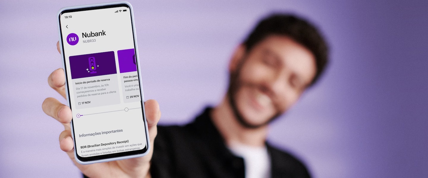 Imagem de um homem com o braço esticado para frente, segurando um celular com a tela voltada para a câmera. Ele está borrado no fundo da imagem. No primeiro plano, o celular mostra a tela de IPO do Nubank