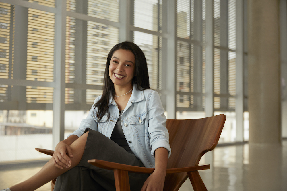 Uma mulher sentada com a perna cruzada, sorrindo para a câmera. Ela usan um vestido cinza e uma jaqueta jeans