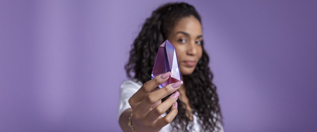 NuSócios: uma mulher segura um pedacinho do Nu (na forma de um cristal roxo) na mão, apontando para a câmera.