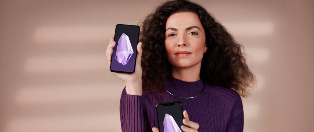 Pedacinho: mulher vestindo uma blusa roxa segura um celular; na tela do celular aparece um cristal roxo, o pedacinho do Nubank.