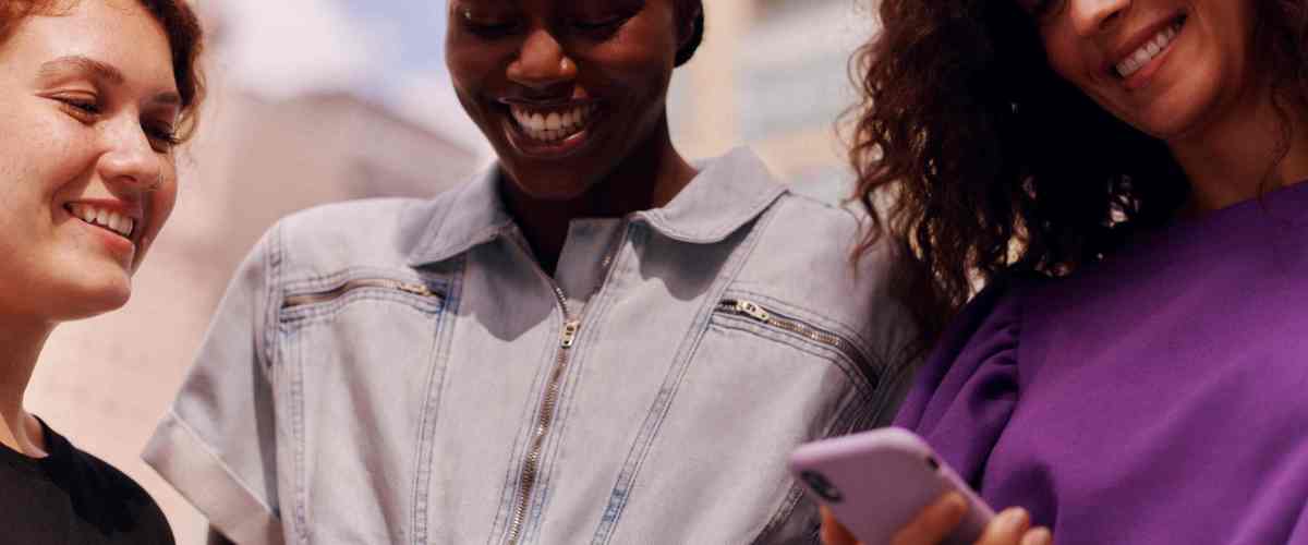 Na imagem aparecem tries mulheres jovens, sorrindo, e a da direita está olhando para a tela do celular