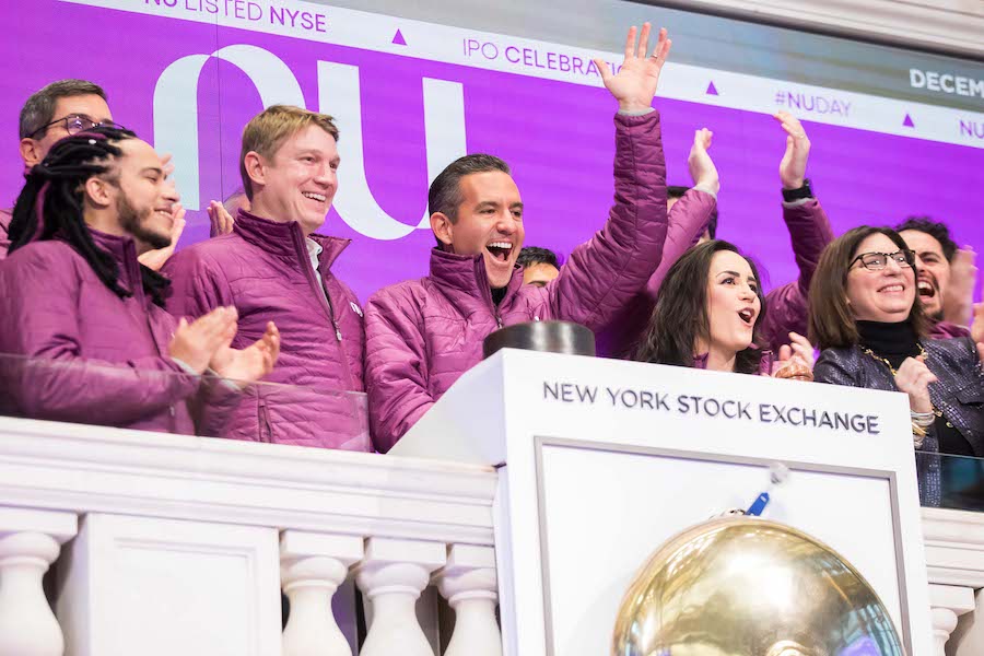Várias pessoas vestidas com jaquetas magenta tocam o sino da NYSE, a Bolsa de Nova York. No centro, tocando o sino, está David Velez, CEO do Nubank.