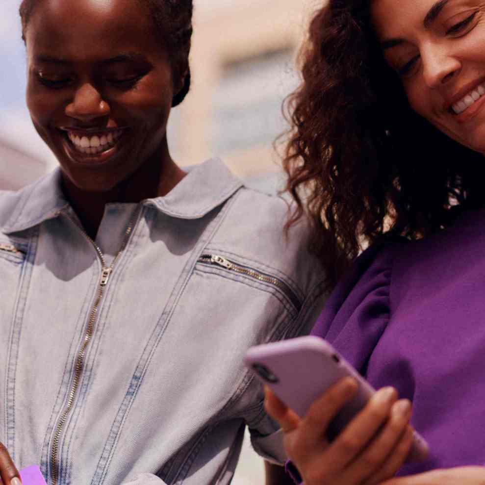 Na imagem aparecem tries mulheres jovens, sorrindo, e a da direita está olhando para a tela do celular