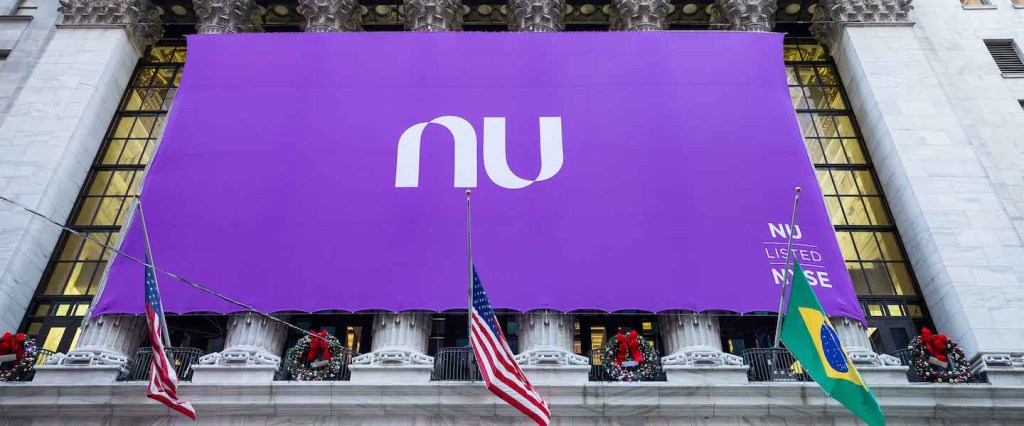 Nubank na Bolsa de Valores: prédio da NYSE, em Nova York, coberto por uma bandeira roxa gigante escrito NU no centro. Logo abaixo estão duas bandeiras dos Estados Unidos e uma do Brasil