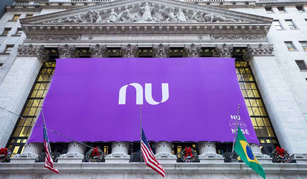 Nubank na Bolsa de Valores: prédio da NYSE, em Nova York, coberto por uma bandeira roxa gigante escrito NU no centro. Logo abaixo estão duas bandeiras dos Estados Unidos e uma do Brasil
