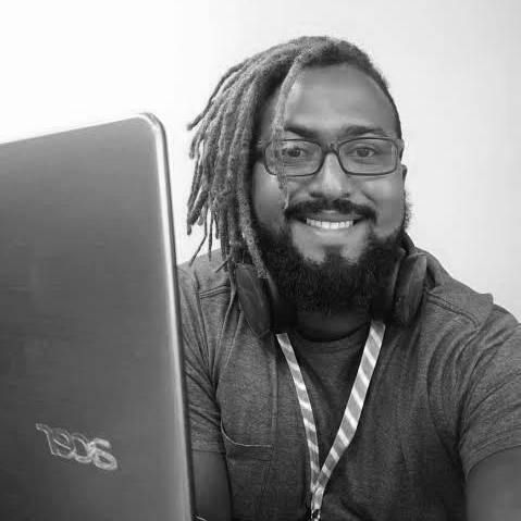 Na imagem aparece Leandro, um homem negro, de barba, usando óculos e cabelo trançado. Ele usa uma camiseta, está com fone de ouvido e também aparece um pedaço do seu laptop à esquerda.