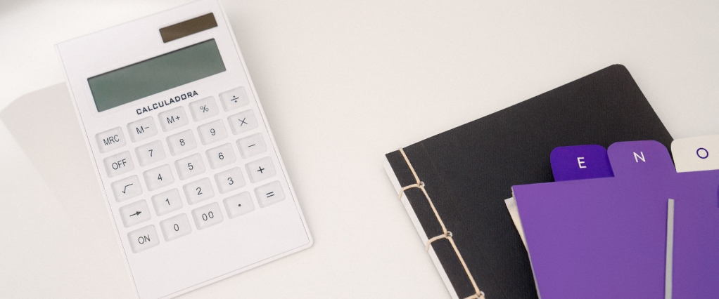 Pis/Pasep 2024: imagem de uma calculadora branca ao lado de um caderno preto e de etiquetas roxas sobre uma mesa de escritório branca.