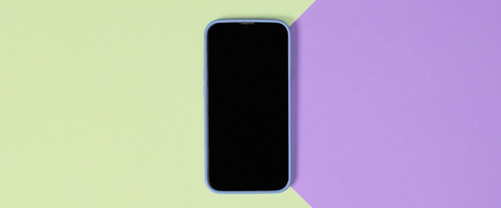 imagem de um celular com a tela preta no centro de uma superfície de cores verde claro e lilás.