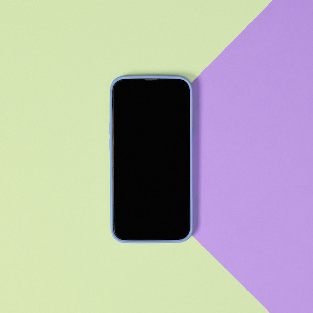imagem de um celular com a tela preta no centro de uma superfície de cores verde claro e lilás.
