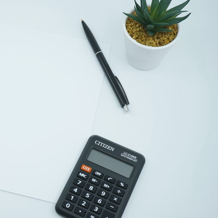 Endividamento e inadimplência: imagem de uma calculadora preta sobre uma mesa branca com folha de papel, clips, caneta e vaso de suculenta simbolizando contabilidade de dívidas. Foto: @mediamodifier/Unsplash